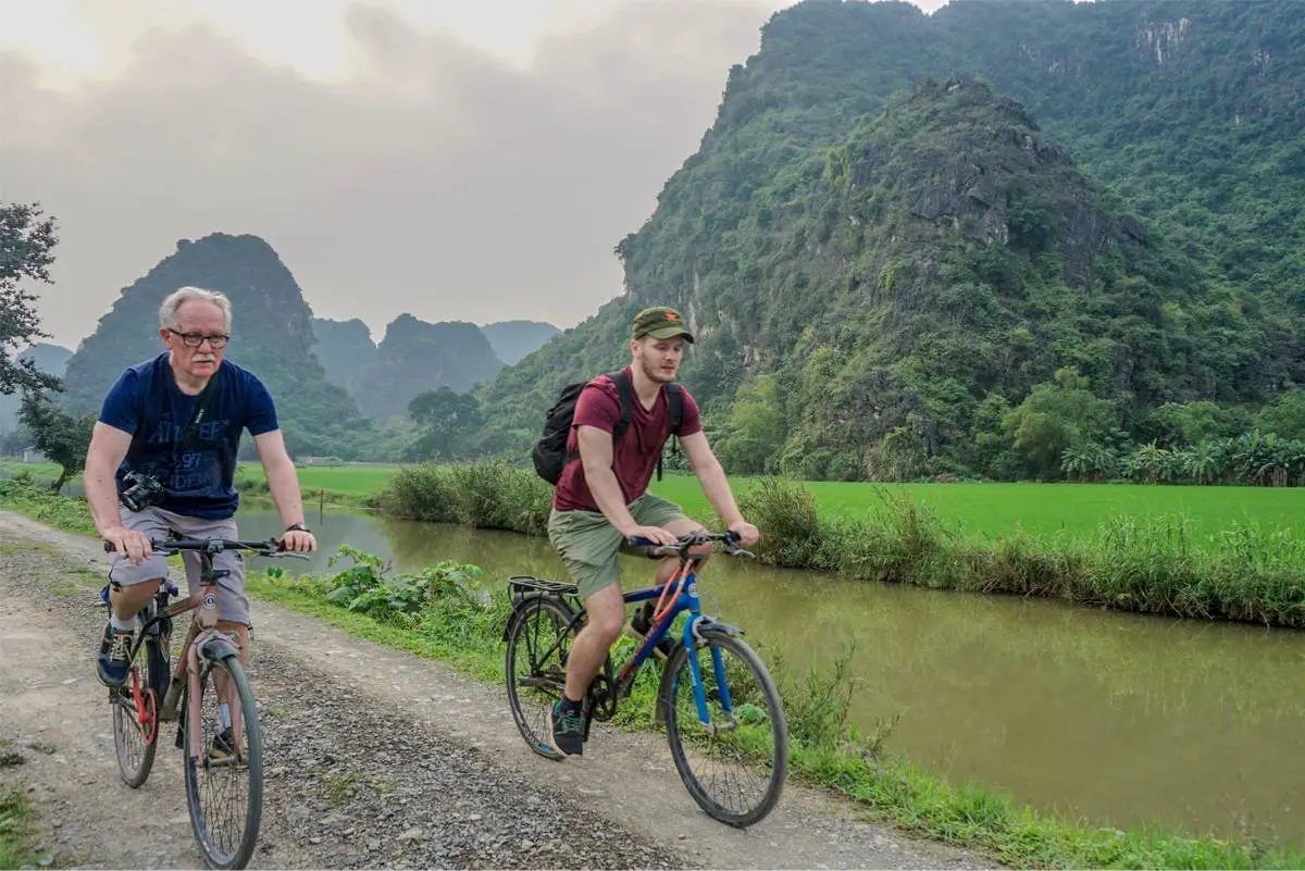 Cyclists riding a rice paddy road beneath limestone karst towers near Hoa Lu, the natural fortress walls of Vietnam's first capital